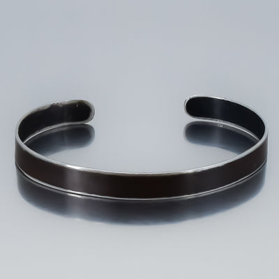 The Marvin - Oldsmobile is a minimalist, open-ended metal cuff bracelet with a smooth dark band inspired by vintage car parts and shiny silver edges, showcased on a neutral gray background.
