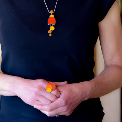 A person in a black shirt showcases the Dotty - Ford & Jeep necklace and matching stainless steel ring, both featuring orange and yellow circular designs. Their clasped hands at waist level draw attention to the vibrant jewelry.