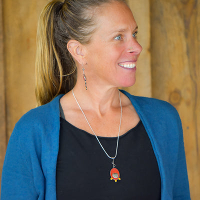 A smiling woman with a ponytail wears a blue cardigan and black top, her Fannie - Mercury & Ford jewelry—featuring a long silver necklace with a red pendant and matching dangling earrings—stands out against the wooden background.