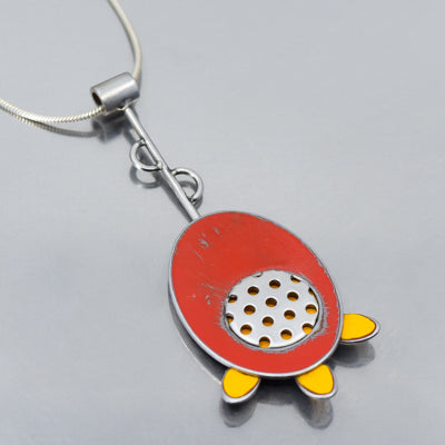 The Fannie - Mercury & Ford pendant features a red oval rocket with yellow fins, crafted from vintage car parts with original paint, silver dotted detail, and hangs from a silver chain on a reflective surface.
