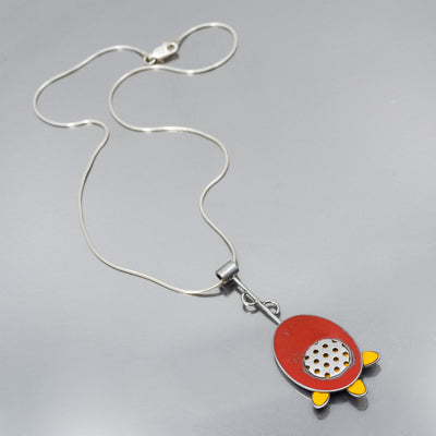 The Fannie - Mercury & Ford is a striking silver necklace with a turtle pendant, featuring a red oval body, yellow feet, and a central silver circle—jewelry inspired by vintage car parts and displayed on a plain light gray background.