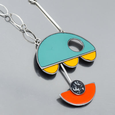 Leona - Chevy & Ford is a modern, abstract pendant inspired by vintage car parts, featuring a turquoise semicircle with cutout, yellow scalloped edge, orange semicircle, and silver chain on a gray background.