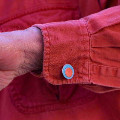A close-up of a person’s arm wearing a red long-sleeve shirt with the Melvin - Ford cufflink—a light blue and orange circle crafted from vintage car parts—on the cuff. The blurred background features the same red shirt fabric.