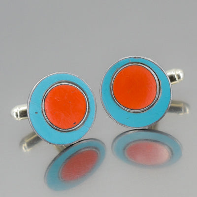 A pair of Melvin - Ford round cufflinks with a bright orange center and turquoise blue outer ring, inspired by vintage car parts, displayed on a reflective surface.