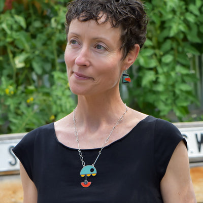 A woman with short curly hair in a black top stands outdoors, showcasing the Leona - Chevy & Ford jewelry made from vintage car parts with blue, gray, and orange shapes. Green foliage and a street sign are visible behind her.