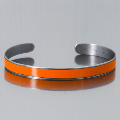 The Dolores - AMC Rambler is a silver metal cuff bracelet featuring a vivid orange enamel strip, inspired by vintage car parts and original paint, shown on a reflective gray background.