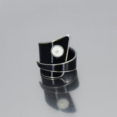 The Randall - GMC & Ford is a modern black asymmetrical wrap ring with a shiny finish and a small circular silver accent, inspired by vintage car parts, shown on a reflective gray surface.