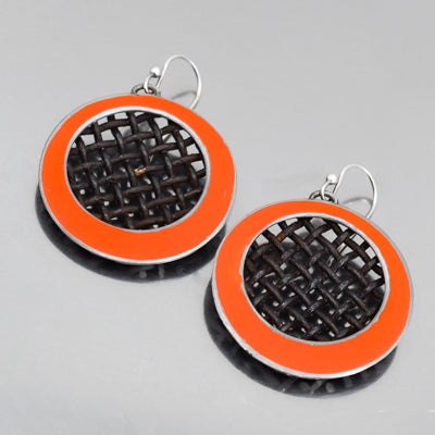 The Dorthy -Ford pickup earrings feature round black woven centers and vibrant orange borders, with metal hooks, evoking vintage car parts with original paint, displayed on a reflective gray surface.