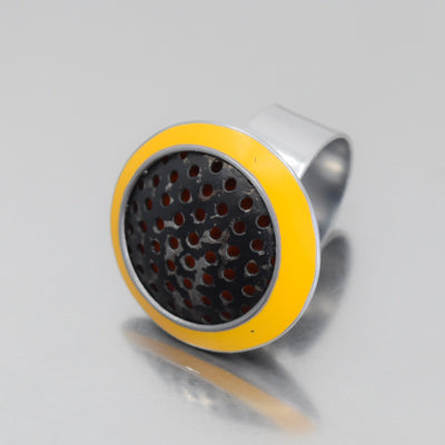 The Mable - Ford ring features a large, round top with a black perforated center and bold yellow border, giving it a vintage car part aesthetic. Shown on a reflective gray surface, this modern silver piece stands out.