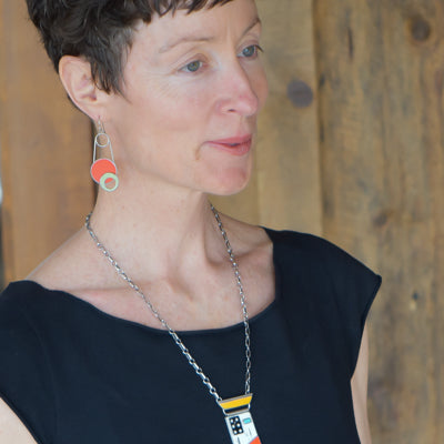 A woman with short brown hair wears the Sallie - Ford & Plymouth black top, geometric earrings, and a matching pendant necklace made from vintage car parts, standing before a wooden background.