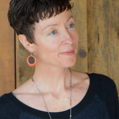 A woman with short curly brown hair in a black top and the Dorthy -Ford pickup jewelry—crafted from vintage car parts, featuring a long silver necklace and large round earrings—stands before a wooden wall, gazing softly to the side.