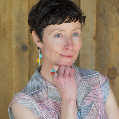 A person with short, wavy brown hair and blue eyes models the Myrtle - AMC, Ford, Pontiac geometric jewelry made from vintage car parts, wearing a sleeveless top and posing against a wooden background.