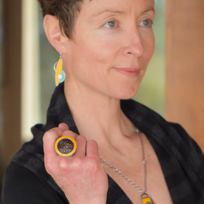 A person in a black top models the Mable - Ford: a yellow honeycomb-pattern ring, matching yellow earrings, and a vintage car part necklace, standing indoors by a wooden structure.