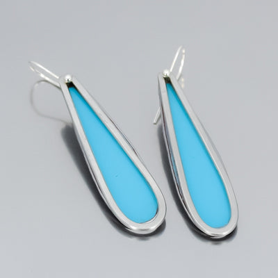 A pair of teardrop-shaped earrings, crafted from vintage Ford Thunderbird car parts, feature bright turquoise centers outlined in shiny stainless steel metal and are displayed on a light gray background.