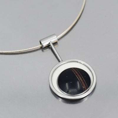 Introducing Maggie - Ford: a silver necklace with a round pendant, black stone center, and white border—this unique piece draws inspiration from vintage car parts and original paint, elegantly displayed on a reflective surface.