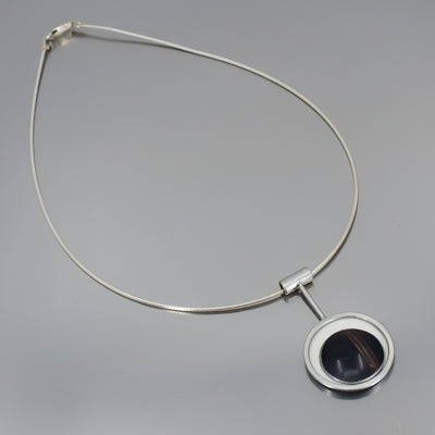 Maggie - Ford: A silver necklace with a circular pendant, featuring a black stone center and white border inspired by vintage car parts with original paint, shown on a light gray background.