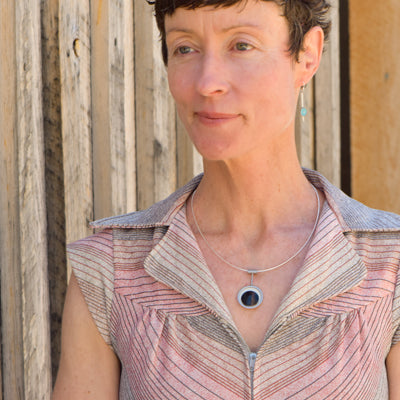 A woman with short brown hair, wearing the Maggie - Ford necklace—a circular pendant crafted from vintage car parts—stands before a wooden wall, looking slightly to the side and smiling gently.