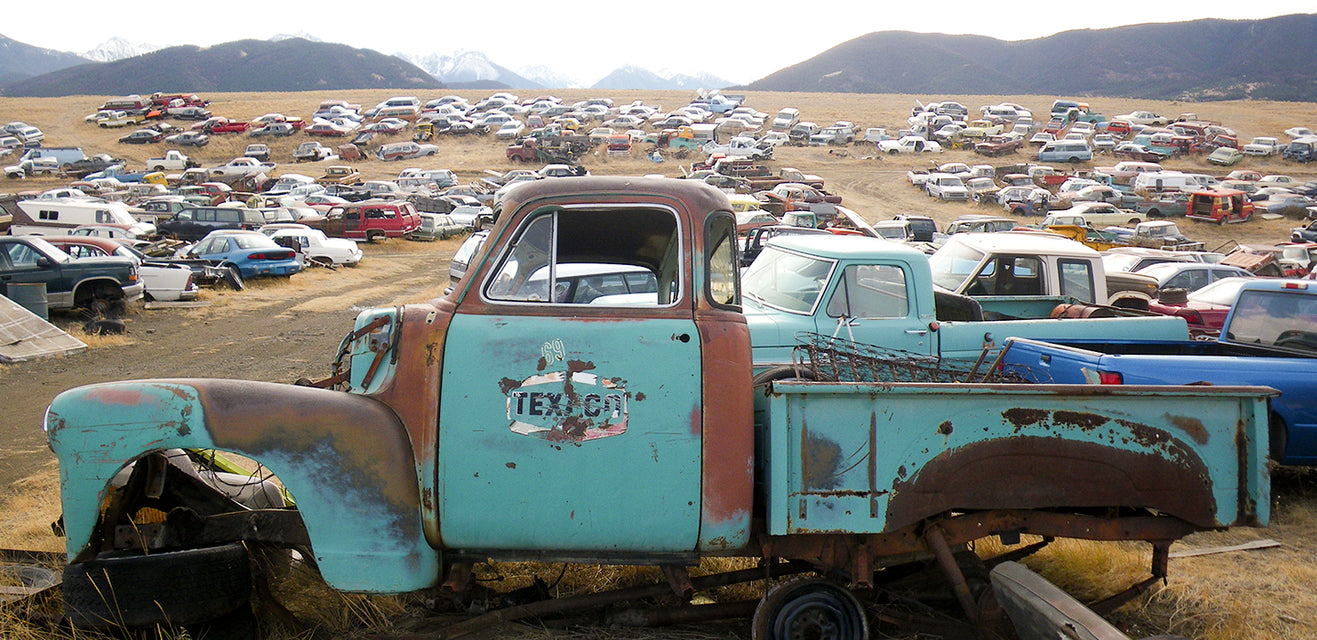 A rusted, turquoise pickup truck with TEXACO on the door sits in the foreground of a large junkyard filled with old cars and original paint, set in a grassy field with mountains in the background.