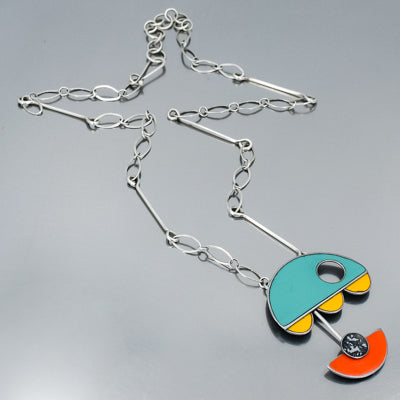 The Leona - Chevy & Ford necklace features a silver chain and an abstract pendant with teal, orange, and yellow shapes, capturing the bold modern style of vintage car jewelry. Its geometric design stands out beautifully on a gray background.