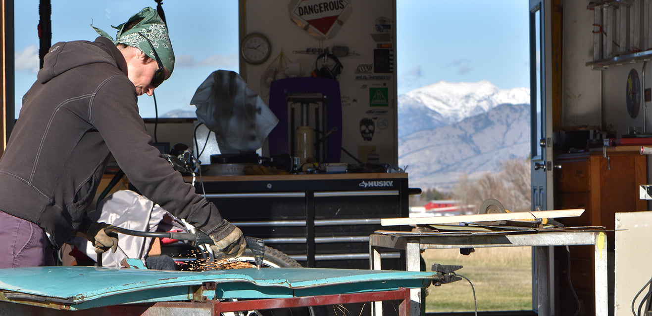 A person wearing a green bandana and gloves uses tools to work on vintage car parts in a workshop, with mountains and blue sky visible through an open door in the background.