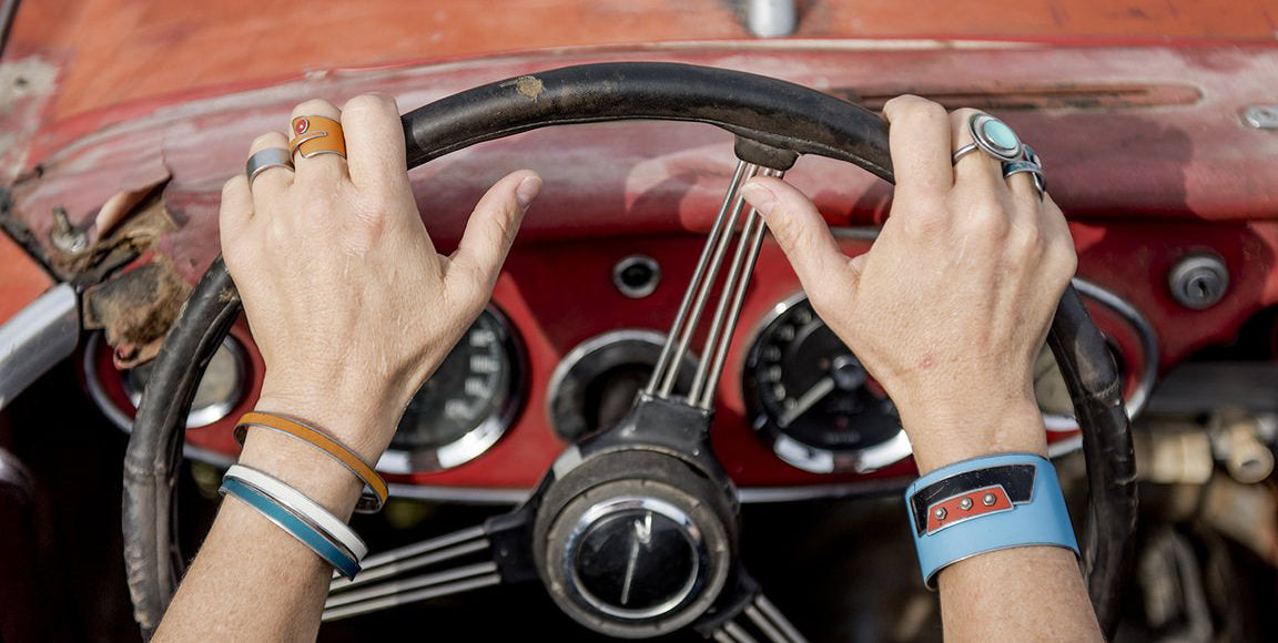 Close-up of two hands adorned with jewelry gripping the steering wheel of an old, red, vintage car; dashboard, gauges, and original paint visible in the background.