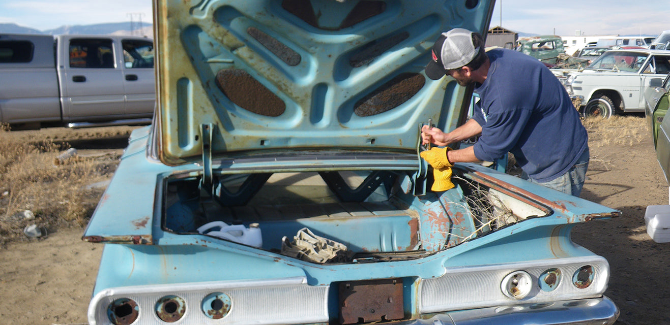 A person in a cap is working on the trunk of a vintage blue car with original paint and a rusty exterior in a junkyard, surrounded by other old vehicles and dry grass.