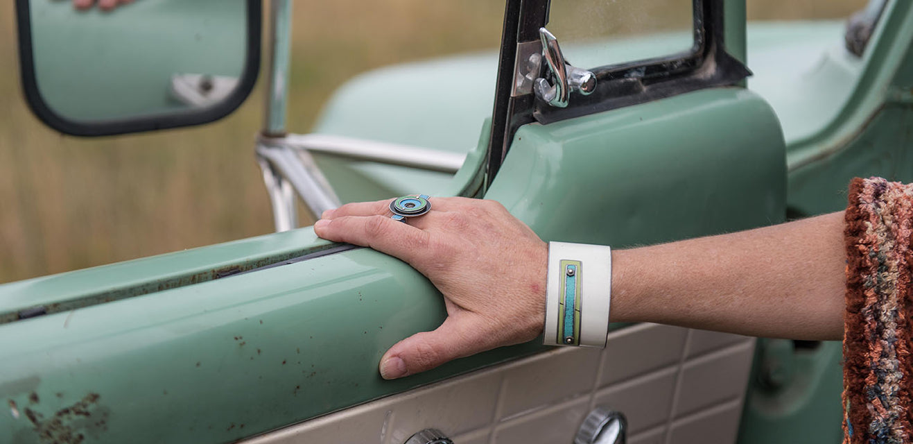 A person rests their hand, adorned with eye-catching jewelry, on the open door of a vintage mint-green truck. The original paint gleams as the truck’s mirror and window frame a grassy field in the background.