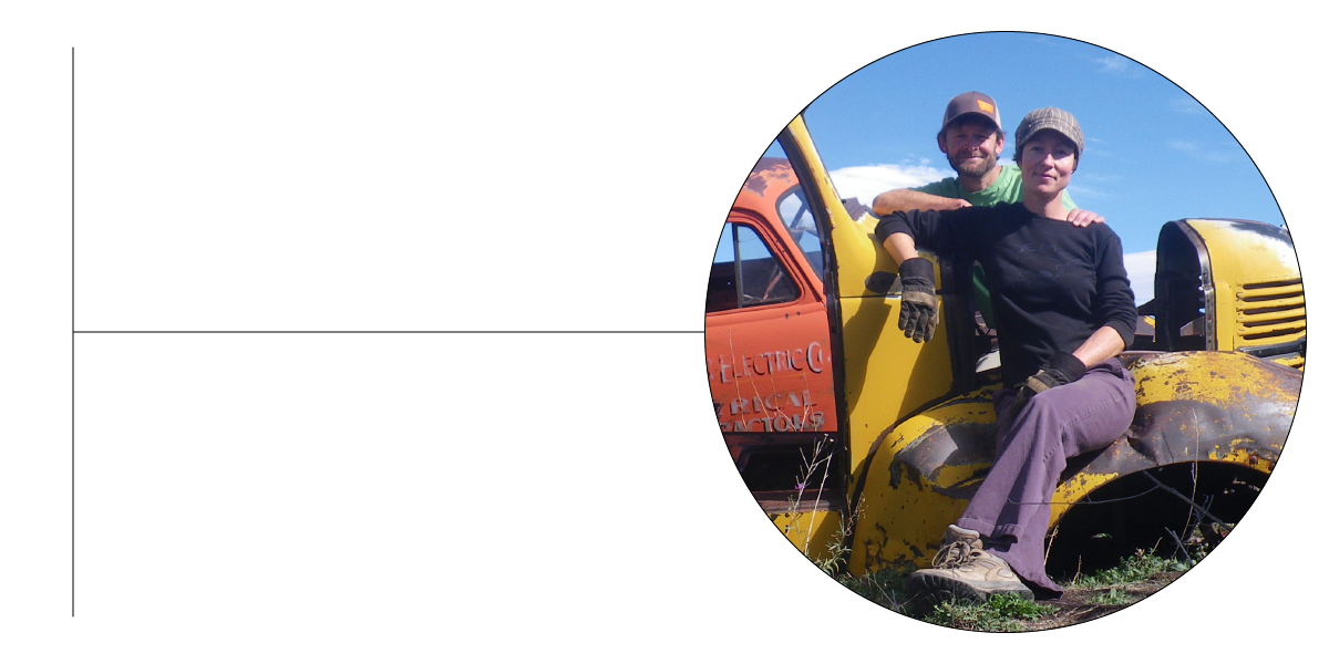 A smiling man and woman wearing gloves pose by a rusty yellow and orange vintage truck with original paint outdoors under a blue sky.