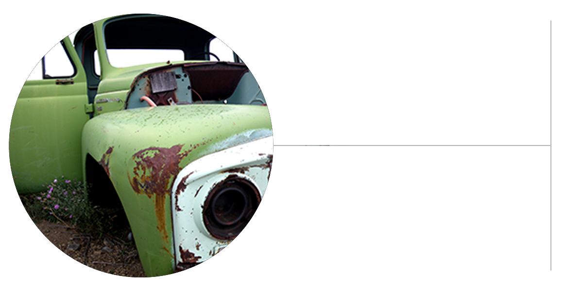 A close-up of an old, rusted green car with original paint, a damaged hood, and missing parts, shown outdoors among grass and wildflowers. The car is partially framed in a circular crop.