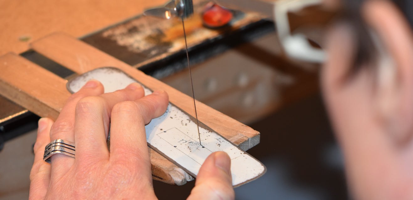 A person uses a jeweler’s saw to cut a shape out of a flat piece of metal, perhaps repurposed from vintage car parts, focusing closely on precise work at a wooden workbench.