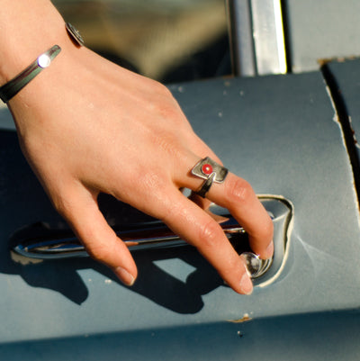 A hand wearing Randall - GMC & Ford jewelry—a silver ring with a red stone and a bracelet—grasps the handle of a vintage blue car door, its original paint gleaming softly.