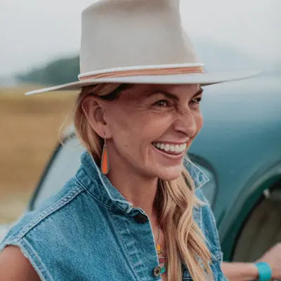 A woman with long blonde hair, in a wide-brimmed hat, denim vest, and orange jewelry, smiles brightly while standing outdoors next to Willie—the AMC Rambler with its original paint.