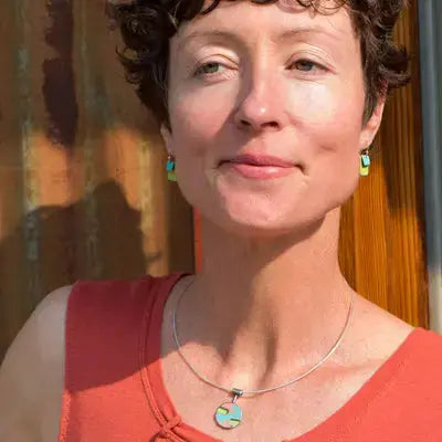 A woman with short curly hair wears a sleeveless orange top and the Etta - Ford: unique jewelry made from vintage car parts with original paint, standing in sunlight against a wooden and metal background.