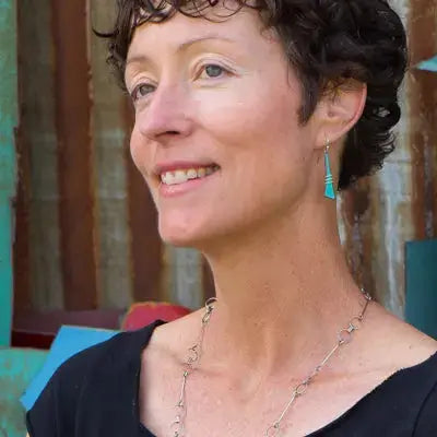 A woman with short curly brown hair, wearing a black top, turquoise earrings, and a silver necklace stands smiling near Maxine, a Ford pickup with vintage teal and rusty car parts in the background.