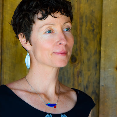 A woman with short curly dark hair, wearing a black top, silver leaf earrings, and the Eloise - Dodge pickup necklace made from vintage car parts with original paint, stands before a rustic wooden background.
