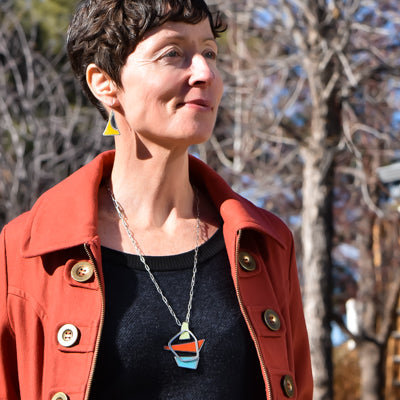 Wearing a rust-colored coat and black top, a woman with short brown hair stands outdoors in sunlight. She showcases the Earnestine - Plymouth & Ford set: geometric earrings and a matching pendant necklace made from vintage car parts.