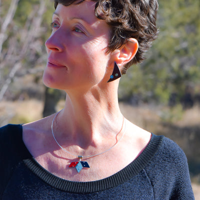 A woman with short brown hair wears the Darcy - Mercury, Dodge, Chevy black top and matching geometric earring and pendant in black, red, and blue. Outdoors near trees and a classic Chevy pickup, she gazes thoughtfully to the side.