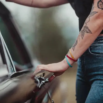 A person with leaf tattoos and jewelry opens the door of Dolores - AMC Rambler. Their colorful bracelets catch the light, jeans visible, while a blurred background reveals hints of original paint on this vintage car.