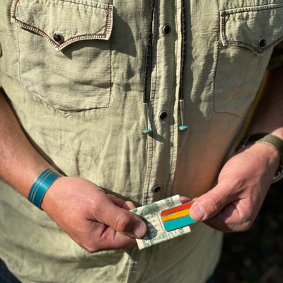 A person in a light green button-up holds a folded dollar with the Ernesto - Dodge Dart money clip, featuring red, yellow, and blue stripes. They wear jewelry from vintage car parts plus a teal wristband and watch.