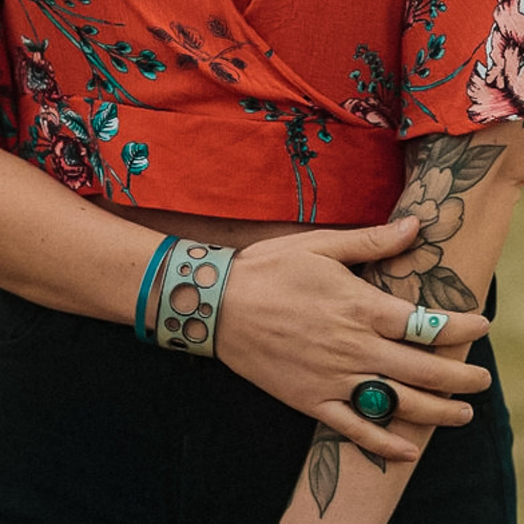 With floral tattoos and chunky rings and bracelets handmade from vintage Ford & Oldsmobile car parts, the person crosses their arms in a red floral top, showing only their tattooed torso and jewelry-adorned arms.