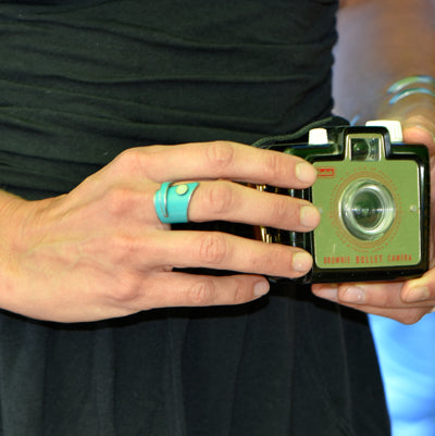 A person wearing a turquoise ring holds the Laurence - Oldsmobile & Plymouth vintage black and silver camera with both hands against a dark background, highlighting the allure of jewelry and the timeless charm of original paint on car parts.