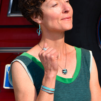 A woman in a green sleeveless top poses confidently wearing matching teal jewelry made from vintage- AMC Rambler car parts, including earrings, necklace, bracelet, and ring, in front of a red  Ford Bronco. Handmade jewelry by Joyride Jewelry.