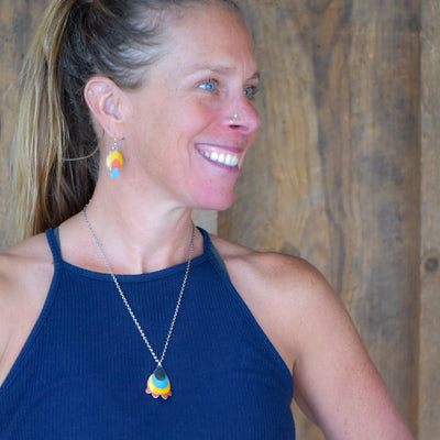 A smiling blonde woman with a ponytail wears the Rhoda - Ford navy blue sleeveless top, colorful drop earrings, and a matching necklace with a sterling silver chain, looking to the side against a wooden background.