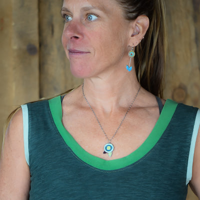 A woman with light skin and blue eyes, wearing a sleeveless dark green top, gazes sideways. Her long hair is pulled back as she models the Fannie - Ford, Dodge, Oldsmobile oxidized sterling silver necklace and dangle earrings with blue accents.