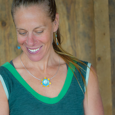 A woman with a nose ring and earrings smiles while looking down, wearing the Edith - Ford & AMC Thunderbird pendant necklace and a green sleeveless top with light green trim, set against a backdrop of wooden planks.