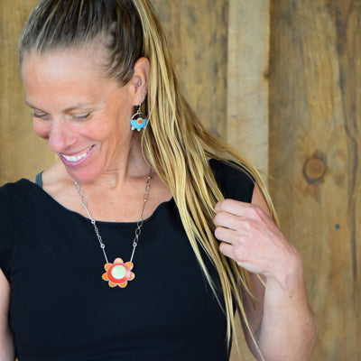 A woman with long blonde hair smiles while looking down, wearing a black top and the Erma - AMC, Mercury, Plymouth flower-shaped pendant on an oxidized sterling silver chain with matching earrings, against a wooden background.