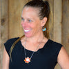 A smiling woman with light brown hair in a ponytail wears a black top and the Erma - AMC, Mercury, Plymouth flower-shaped necklace with matching earrings, standing in front of a wooden background.