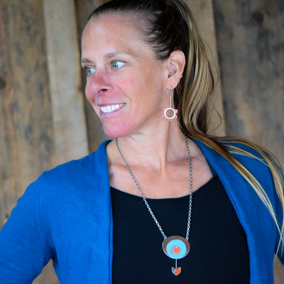 A woman with long blond hair in a ponytail, wearing a blue cardigan and black top, smiles to the side. She wears the Maeve - Ford & Oldsmobile necklace and earrings with circular orange accents in oxidized silver. Rustic wood background.