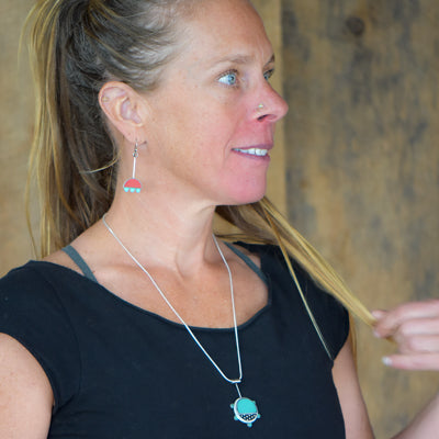 Wearing the Gertrude - Chevy & Ford turquoise pendant with red dangle earrings , a woman in a black shirt stands against a wooden background, gazing to the side and holding her hair.