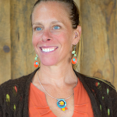 Smiling woman with blue eyes wears the Verna - Ford & Mercury pendant necklace on a sterling silver cable, paired with an orange top, brown textured cardigan, and colorful circular earrings. She stands before a wooden background.